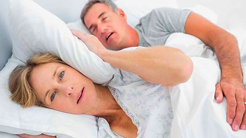 Man snoring loudly in bed while partner covers her ears with pillow and looks annoyed