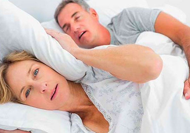 Man snoring loudly in bed while partner covers her ears with pillow and looks annoyed